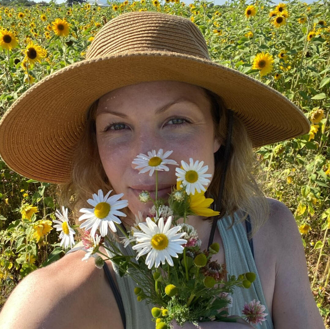 Portrait of Roslyn James in a sunhat, surrounded by wildflowers. She holds a bouquet near her face and looks happy